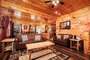 A cozy cabin interior with wooden walls, a ceiling fan, a dining area, and a kitchen.