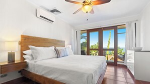 A bedroom with a wooden bed, a nightstand, a ceiling fan, and a view of the outdoors through a glass door. - Ocean View 4 Bedroom Villa Steps to Beach & Resort Amenities (Guanacaste Province)