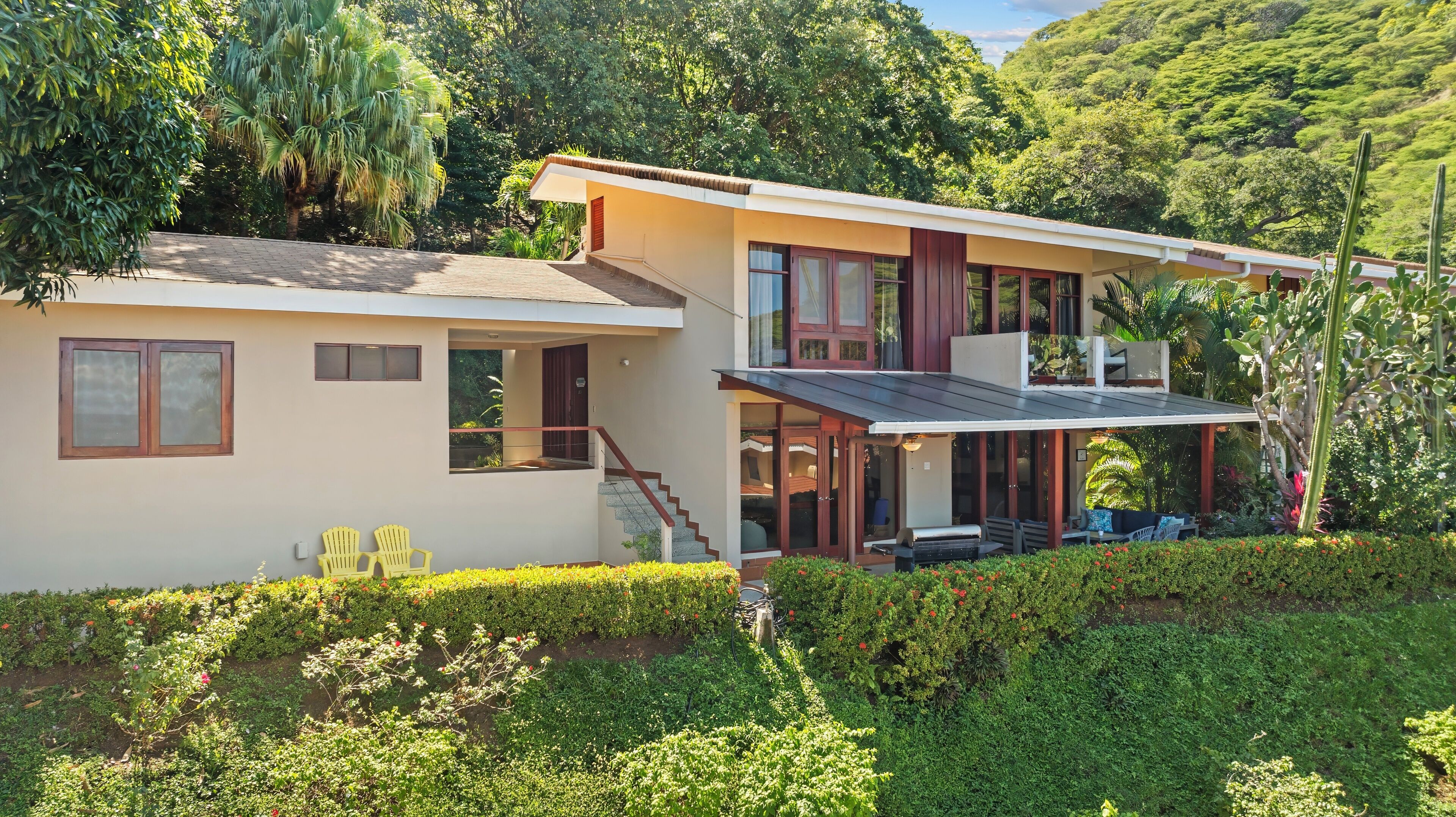 Une maison à deux étages dotée d’un balcon, entourée d’une végétation luxuriante et d’arbres.