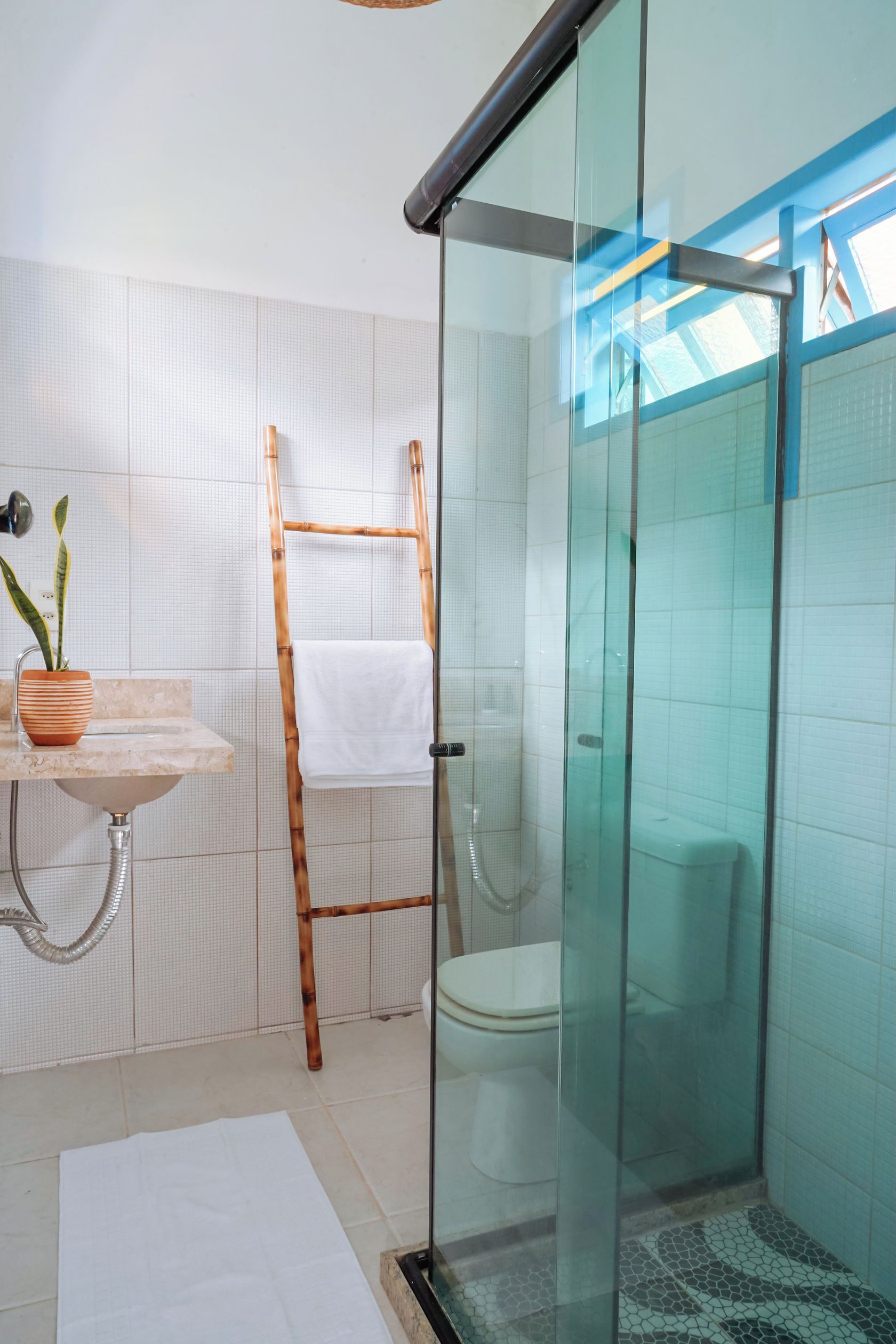 Un baño con cabina de ducha de vidrio, una escalera de madera, un lavamanos con un florero de plantas y una toalla blanca colgada en la pared.