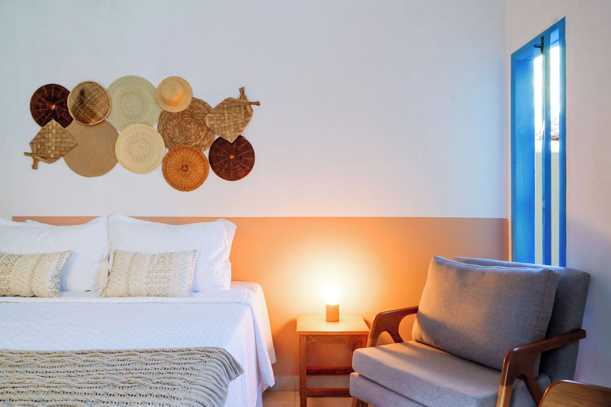 A neatly made bed with pillows, a bedside table with a lamp, a chair, and a wall with decorative items.