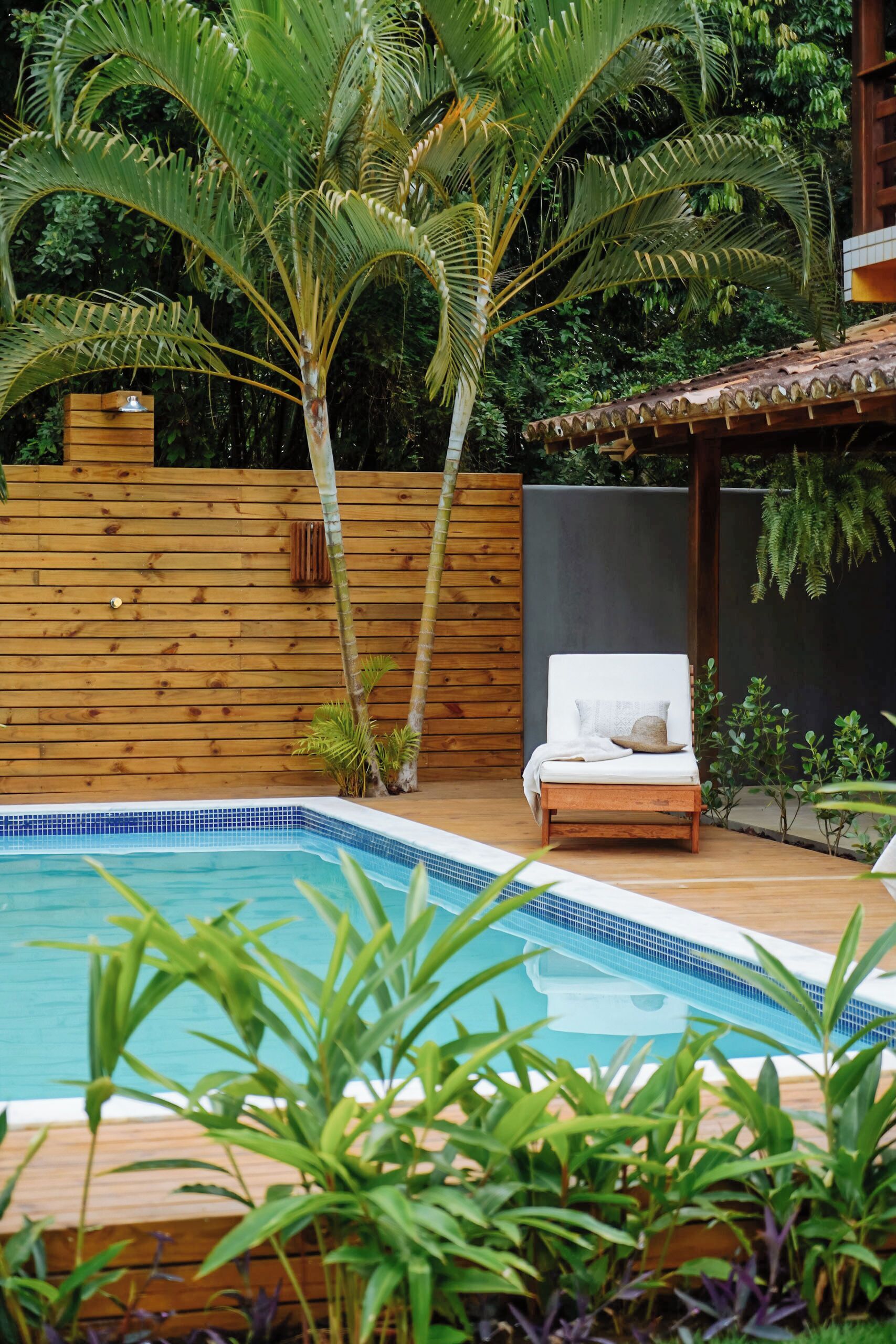 A pool with a wooden deck, a lounge chair, and tropical plants.