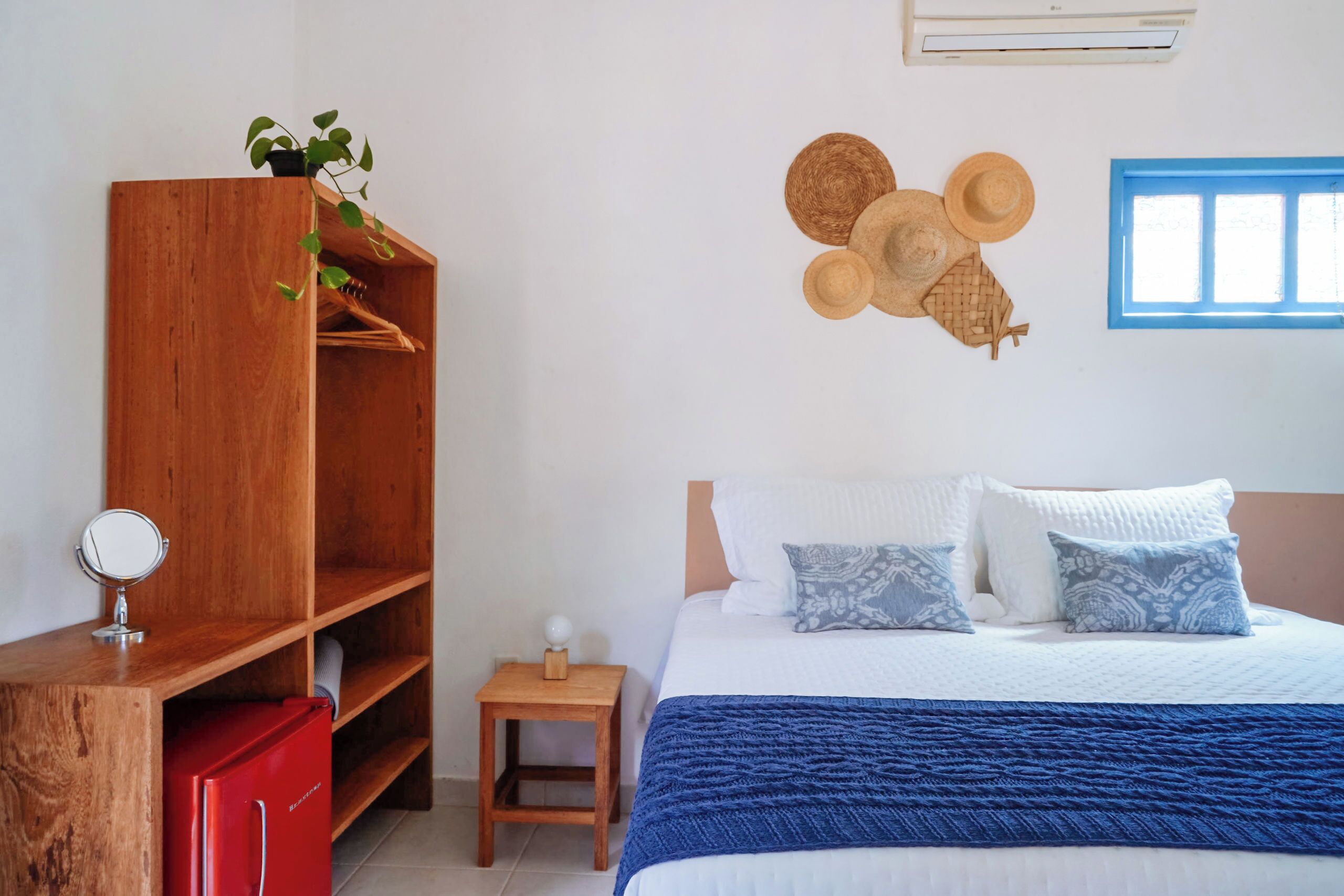 A bedroom with a bed, a wooden wardrobe, a small wooden nightstand, a red refrigerator, and a wall-mounted air conditioner.