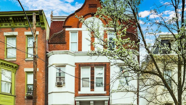 Un bâtiment à deux étages avec une porte rouge et des moulures blanches, une voiture garée devant, et un arbre aux fleurs blanches.