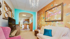 Un salon avec un fauteuil rose, un canapé blanc orné d’un coussin bleu, un meuble en bois et un téléviseur. Un lustre est suspendu au plafond.