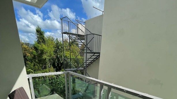 Un balcón con dos sillas de mimbre y una mesita, barandilla de vidrio y vistas a un área verde con árboles y un edificio al fondo.