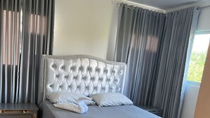 Un dormitorio con cabecera capitonada, una mesita de noche y una ventana con cortinas.