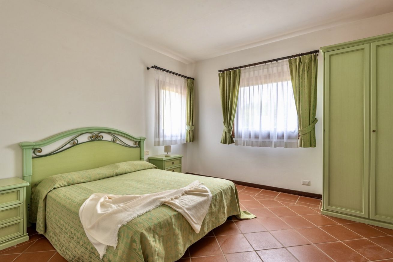 Un dormitorio con una cama verde, dos mesitas de noche, un armario y dos ventanas con cortinas.