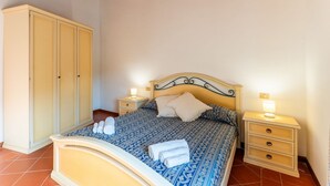 A bedroom with a bed, nightstands, a wardrobe, and a tiled floor. - 60 sq m - Living room with kitchenette and double sofa bed, double bedroom, bathroom with bidet and shower or bath, access to communal pool. <br>Ground floor or first floor with furnished veranda or balcony.<br>Equipped with:<br>- tv <br>- washing machin (Stintino)