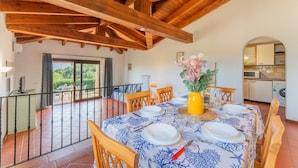 A dining area with a table set for four, wooden beams overhead, a view to the outside, and a kitchen in the background.
