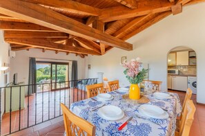 Zona pranzo con tavolo apparecchiato per quattro, travi in legno a soffitto, vista all'esterno e cucina sullo sfondo.