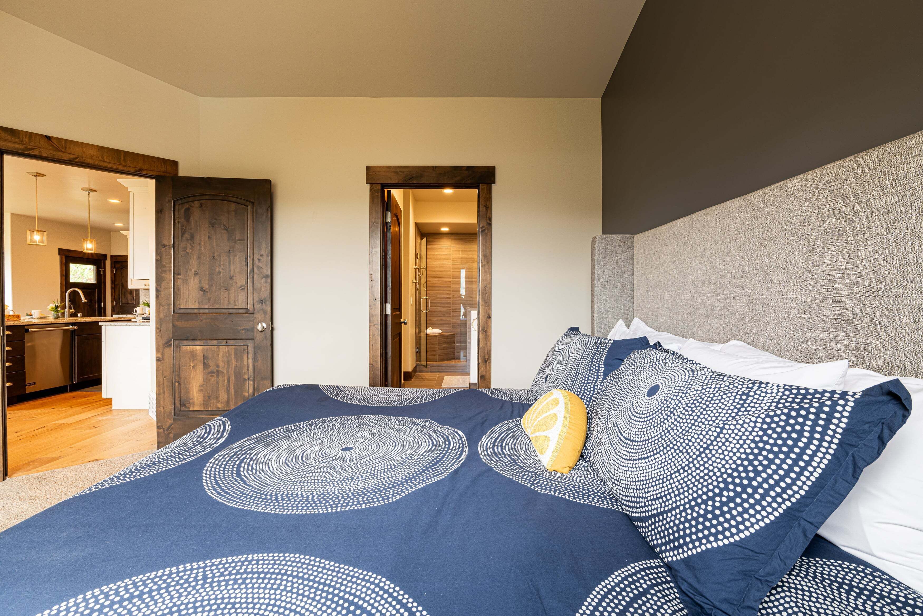 A bedroom with a large bed, a wooden door, and a view into the kitchen.