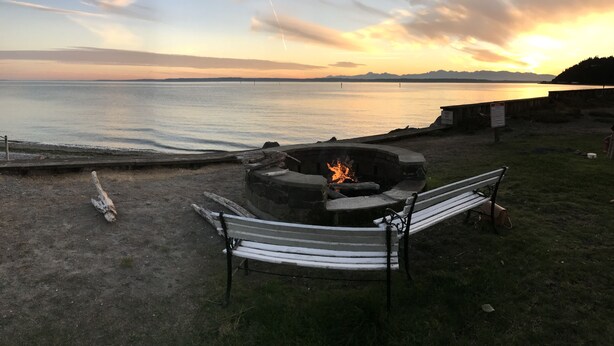 Un décor de plage avec un foyer et un banc, donnant sur une mer calme et des montagnes lointaines.