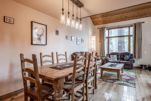 A dining area with a wooden table and chairs, framed artwork on the wall, and a large window with curtains.