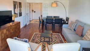 A living room with a sofa, chairs, a coffee table, a TV on a wooden cabinet, a dining table with chairs, and a lamp. - Fabulous Terrace 2 - Checkinhome (Cascais)