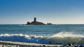 Een rotsachtig strand met golven die op de kust breken en een klein eiland met een toren in de verte.