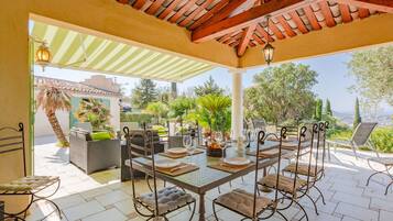 Een terras buiten met een eettafel, omringd door rieten stoelen en een pergola erboven.