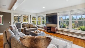 A modern living room with a large TV, comfortable seating, and large windows offering a view of the outdoors.