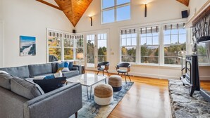 A spacious living room with a high wooden ceiling, large windows, a fireplace, and a cozy seating area.