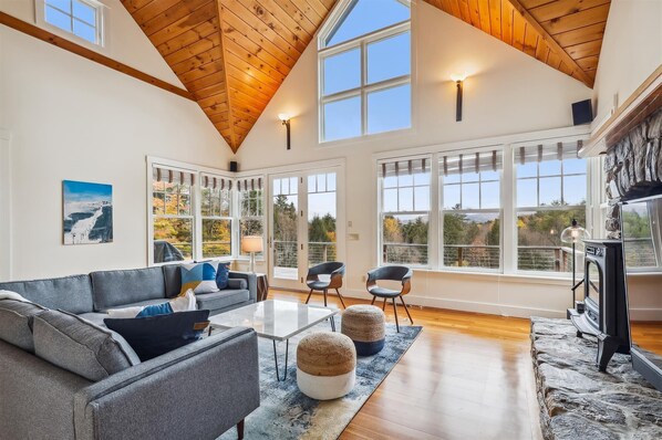 A spacious living room with a high wooden ceiling, large windows, a fireplace, and a cozy seating area.