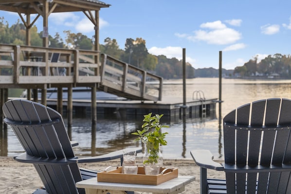 Two Adirondack chairs with a tray table and a glass of water, a vase with green plants, and a wooden dock extending into a body of water.