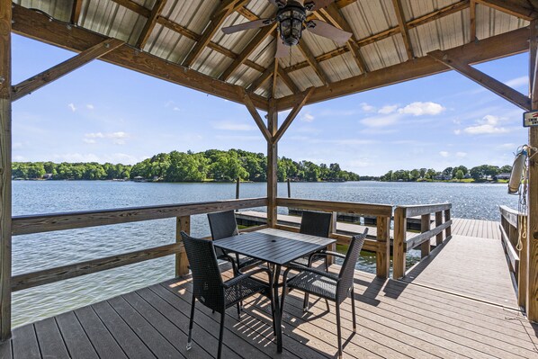 A covered outdoor deck with a table and chairs, overlooking a body of water and a forested area. - Welcome to the Lodge at Greenbay - Sandy Beach & Amazing Sunset Views! (Mooresville)