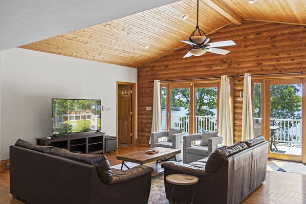 A spacious living room with a wooden ceiling, large windows, and a television on a stand. - Welcome to the Lodge at Greenbay - Sandy Beach & Amazing Sunset Views! (Mooresville)