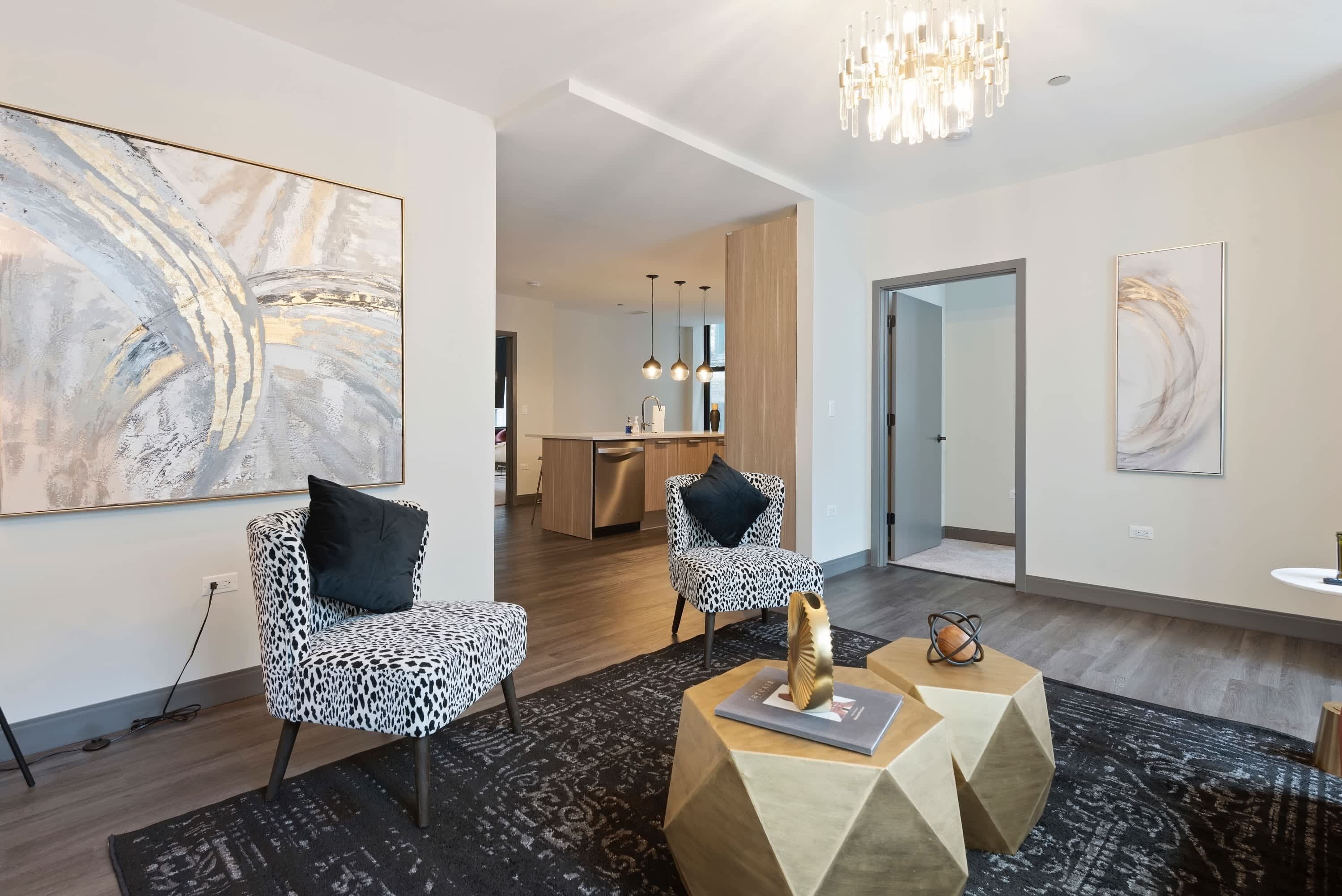 Un salon moderne avec des fauteuils à motifs léopard, des œuvres d’art abstraites, un lustre et une cuisine en arrière-plan.
