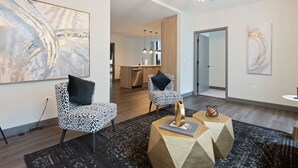 Un salon moderne avec des fauteuils à motifs léopard, des œuvres d’art abstraites, un lustre et une cuisine en arrière-plan.