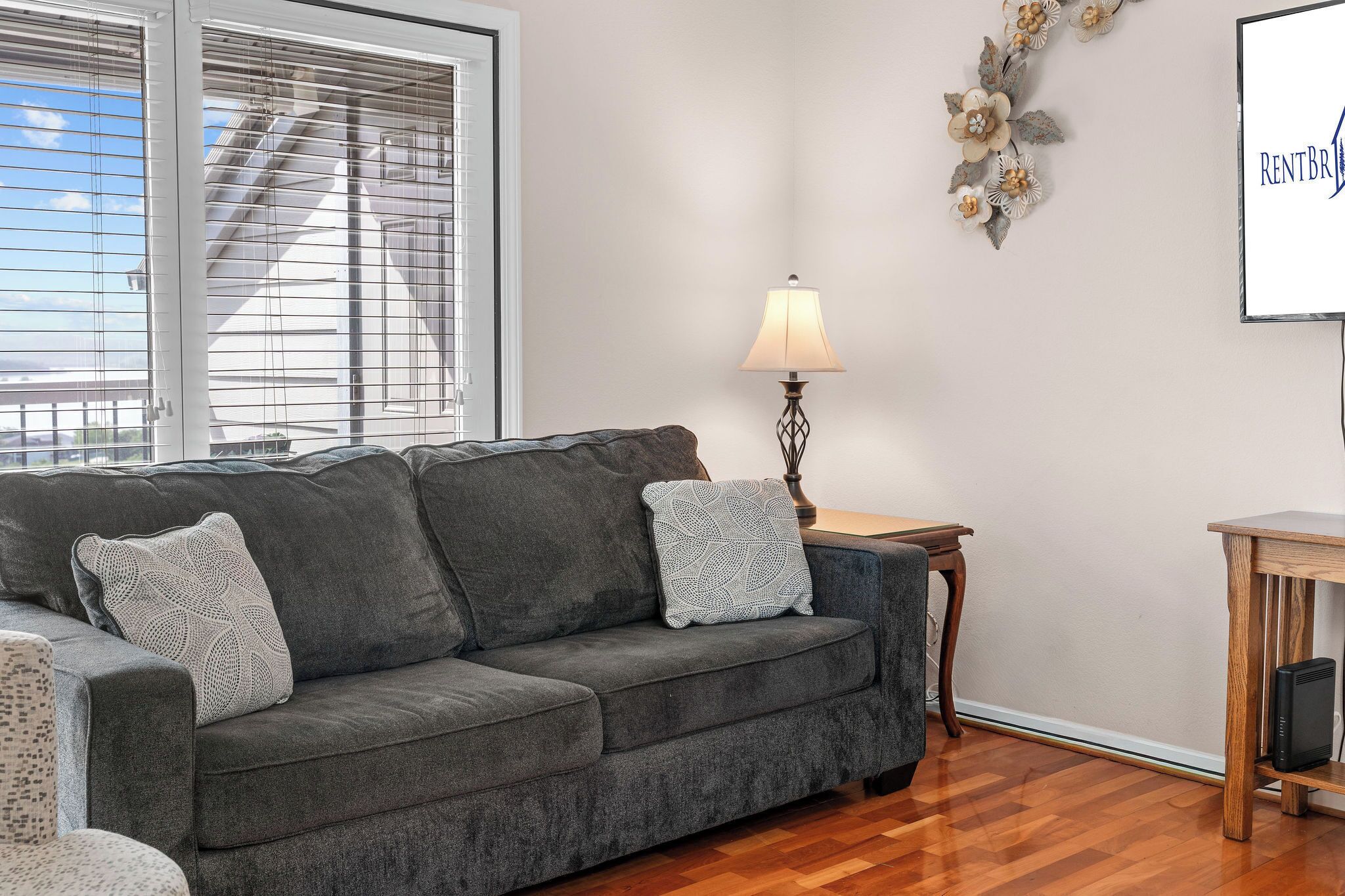 Ein Wohnzimmer mit grauem Sofa, gemusterten Kissen, einem Holzbeistelltisch mit Lampe und einem Wanddekor.