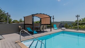 A swimming pool with a wooden deck, lounge chairs, a covered seating area, and a view of the landscape.