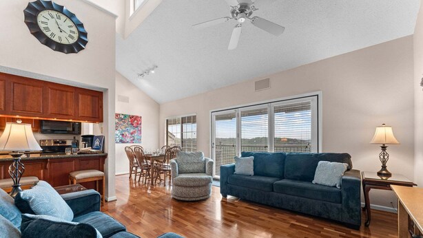 A spacious living room with a blue sofa, a dining area with chairs, a kitchen with wooden cabinets, and a large window with blinds.