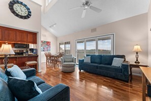 A spacious living room with a blue sofa, a dining area with chairs, a kitchen with wooden cabinets, and a large window with blinds.