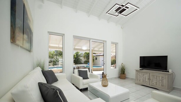 A modern living room with a white sofa, a coffee table, and a TV on a wooden cabinet. Large windows provide views of the outdoors. - Newly Renovated 4BR 4BA Private Pool @ Eagle Beach (Noord)