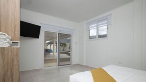A hotel room with a bed, a television mounted on the wall, a sliding glass door leading to a balcony, and a wooden panel on the left side. - Newly Renovated 4BR 4BA Private Pool @ Eagle Beach (Noord)