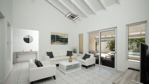 A modern living room with white furniture, a glass coffee table, and a large window overlooking a pool. - Newly Renovated 4BR 4BA Private Pool @ Eagle Beach (Noord)