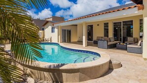 A pool area with a curved edge, outdoor seating, and a covered patio. - Newly Renovated 4BR 4BA Private Pool @ Eagle Beach (Noord)