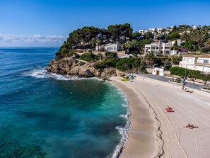 A coastal town with a sandy beach, clear blue sea, and buildings on a hillside.