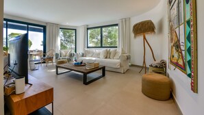 Una sala de estar moderna con sofá, mesa de centro y televisor. Cuenta con ventanas amplias y un cuadro en la pared.