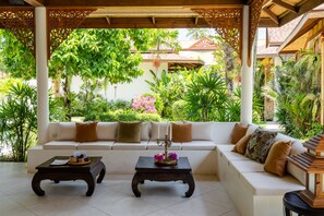 A covered outdoor seating area with a white sofa, two dark wooden tables, and a variety of tropical plants.