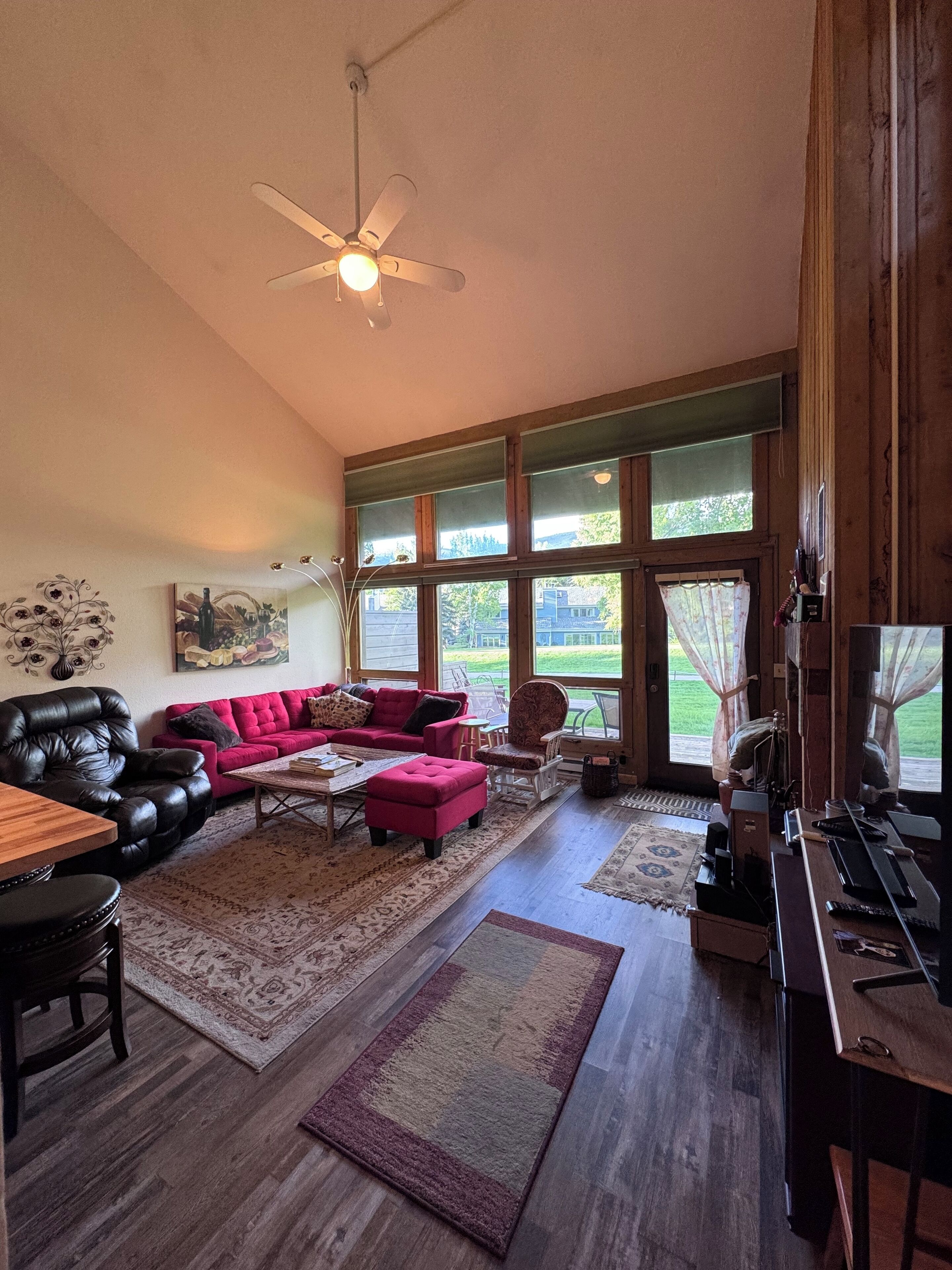 A spacious living room with a leather sofa, a red ottoman, and a wooden coffee table. There is a ceiling fan, large windows with curtains, and a view of the outdoors.