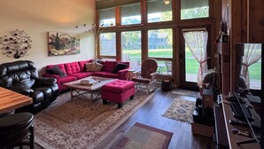 A spacious living room with a leather sofa, a red ottoman, and a wooden coffee table. There is a ceiling fan, large windows with curtains, and a view of the outdoors. - Golf & Ski Townhome in Avon, Colorado (Avon)
