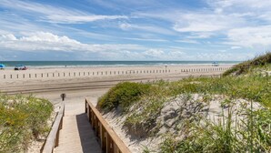 Ein Strand mit einer Holzpromenade, die zum Ufer führt, Sanddünen und einem klaren Himmel.