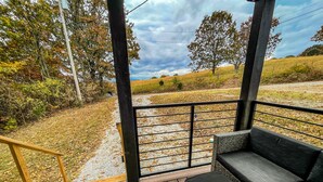 Une terrasse en bois avec une rambarde en métal noir, un canapé en osier et une vue sur un champ verdoyant bordé d’arbres.