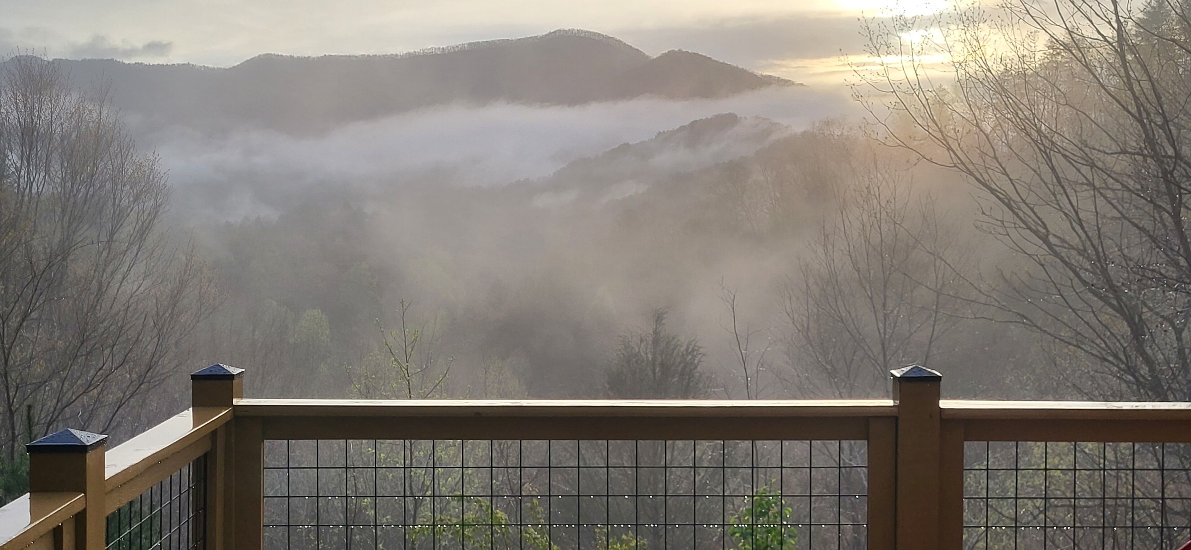 A view from a balcony with a metal railing, overlooking mist-covered mountains and a sunrise.