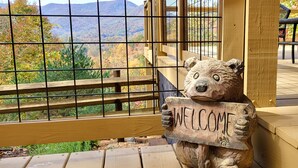 A wooden bear sculpture holding a 'WELCOME' sign, positioned near a window with a metal grid, overlooking a scenic view of mountains and autumn foliage. - Cozy Mountain Chalet with views, private setting! Near WCU and Castle Ladyhawke  (Cullowhee)