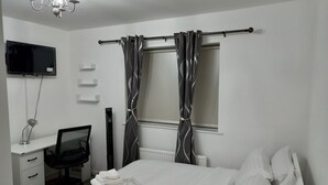 A bedroom with a bed, a desk with a chair, a TV mounted on the wall, and a window with curtains.
