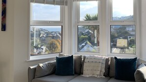 A cozy room with a sofa and three windows offering views of a residential area. - Sea View House in Cornwall Fishing Village (St Austell)
