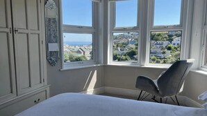 A bedroom with a bed, a chair, and a large window with a view of a town and the sea. - Sea View House in Cornwall Fishing Village (St Austell)