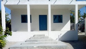 Un bâtiment blanc avec une porte et des fenêtres bleues, un escalier en béton menant à l’entrée et un sol sablonneux.